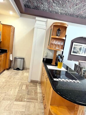 A kitchen with a black countertop, wooden cabinets, a trash bin, and a shelf with glasses. - Puerto Viejo C-2 | Waterfront 3BR on the Malecón (Puerto Peñasco)