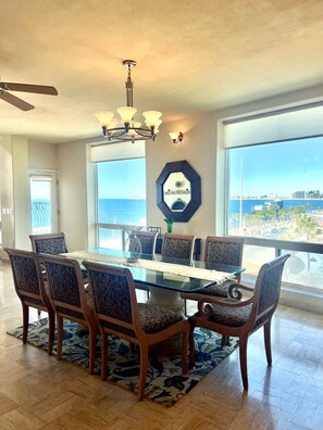 A dining area with a glass table, six chairs, a chandelier, and a view of the ocean. - Puerto Viejo C-3 | Waterfront 3BR on the Malecón (Puerto Peñasco)