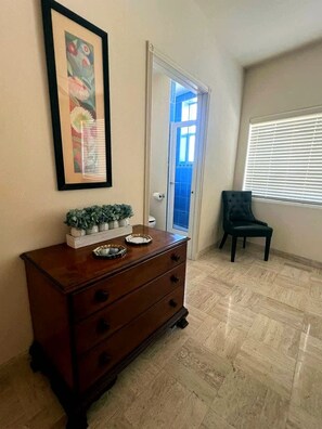 Een kamer met een houten dressoir, een lijst met foto, een deur naar een balkon en een raam met jaloezieën.