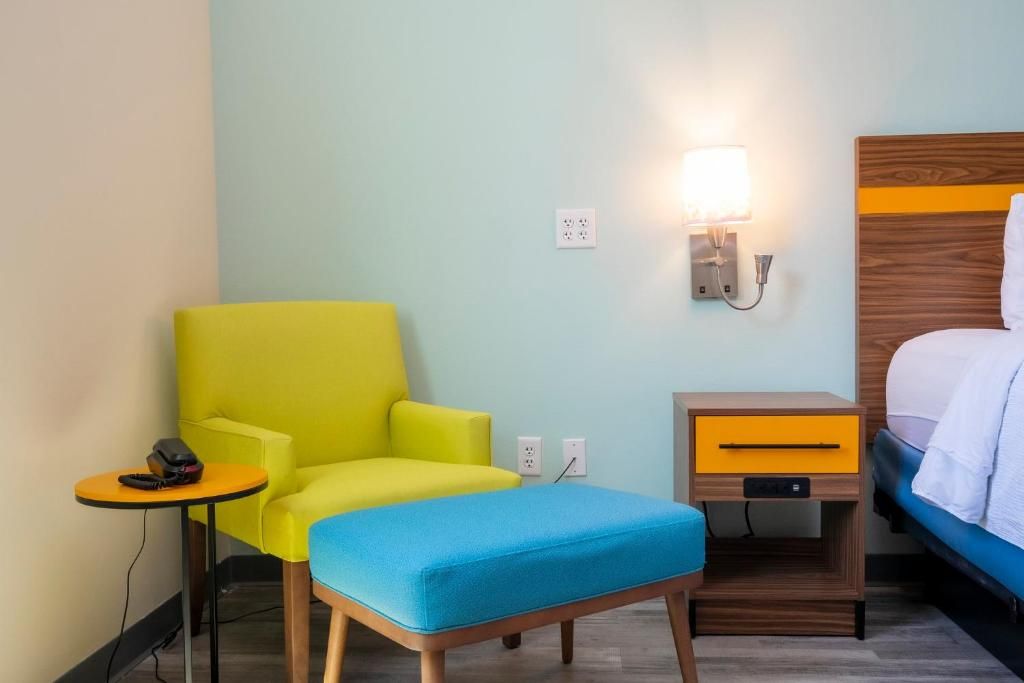 A hotel room with a single bed, a small bedside table with a lamp, a bright yellow armchair, and a blue ottoman.