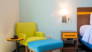 A hotel room with a single bed, a small bedside table with a lamp, a bright yellow armchair, and a blue ottoman.