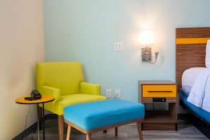 Habitació d'hotel amb un llit individual, una petita taula de nit amb una làmpada, un otomà blau i una butaca groga i brillant.