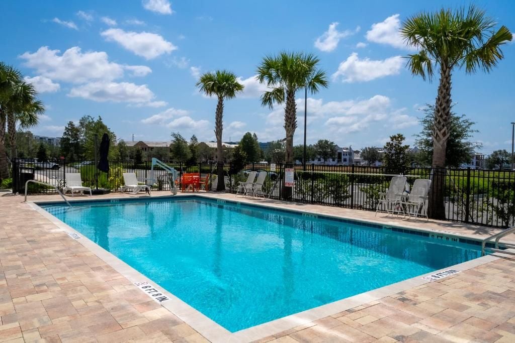 A rectangular swimming pool with a tiled deck, surrounded by a black metal fence, palm trees, and lounge chairs.