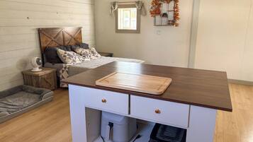 Habitación compacta con cama, mesita de noche y una pequeña zona de kitchenette con tabla de cortar.