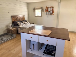 A compact bedroom with a bed, bedside table, and a small kitchenette area with a cutting board.