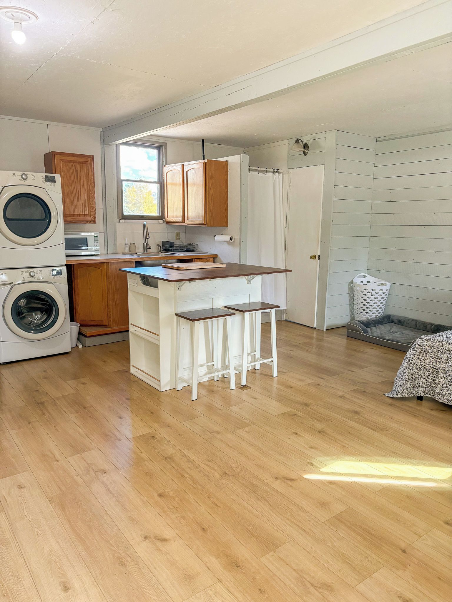 Cocina compacta con lavadora, armarios de madera, una pequeña isla y una cama gris para mascotas.