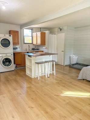 A compact kitchen with a washing machine, wooden cabinets, a small island, and a grey pet bed.