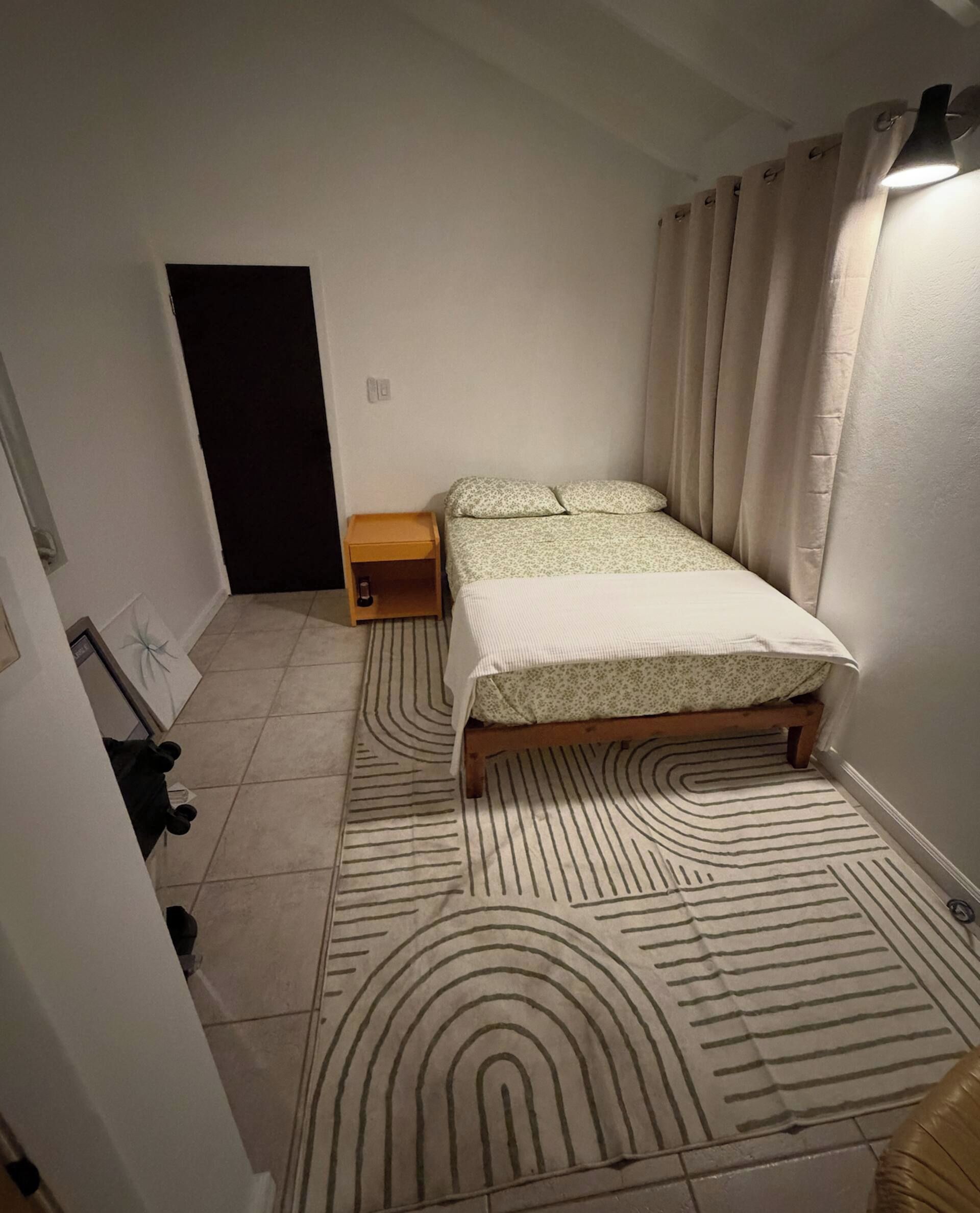 A small bedroom with a bed, a nightstand, a door, and a patterned rug.
