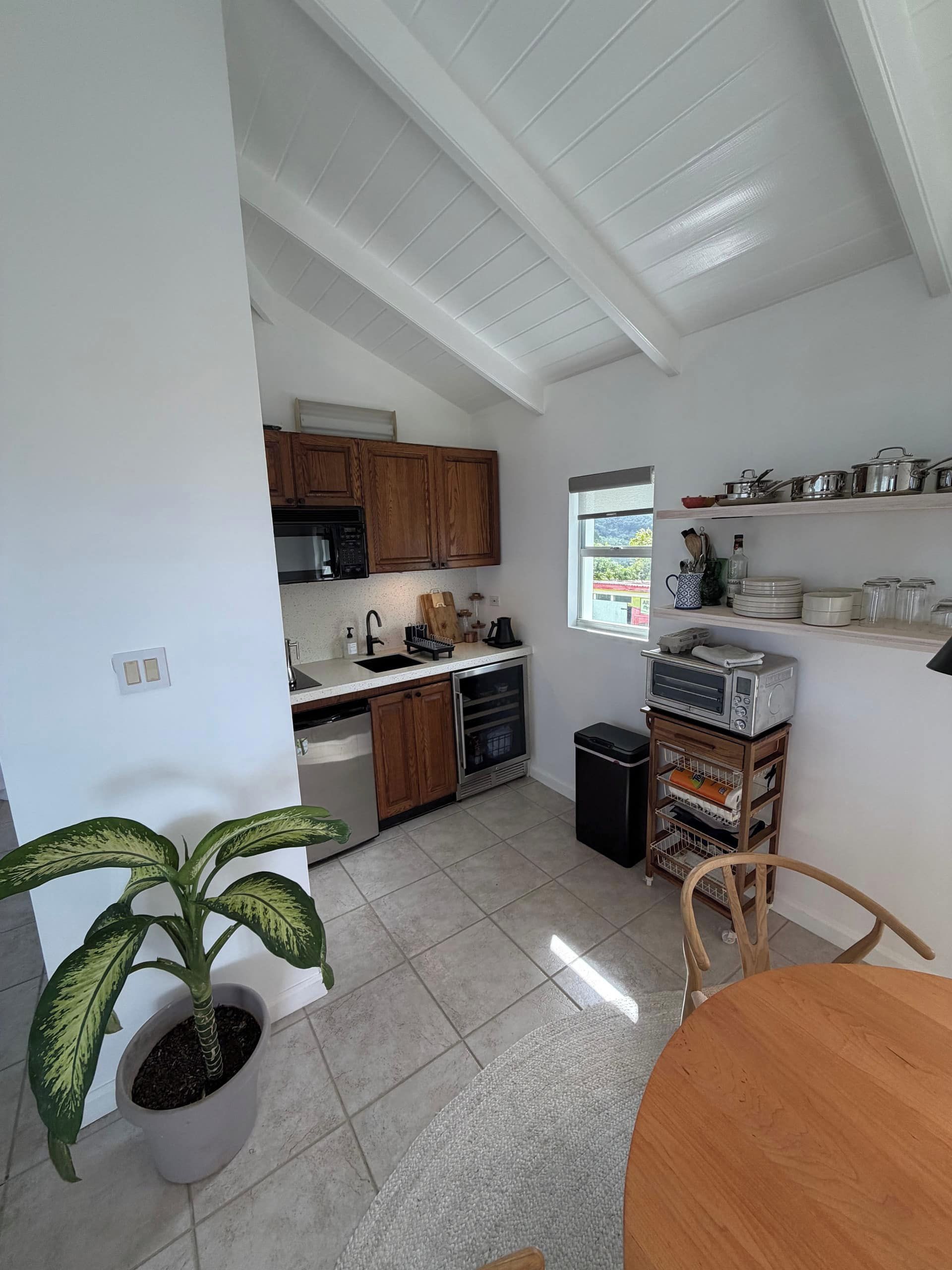 A compact kitchen with wooden cabinets, a microwave, and a sink. There is a potted plant and a dining table with chairs.