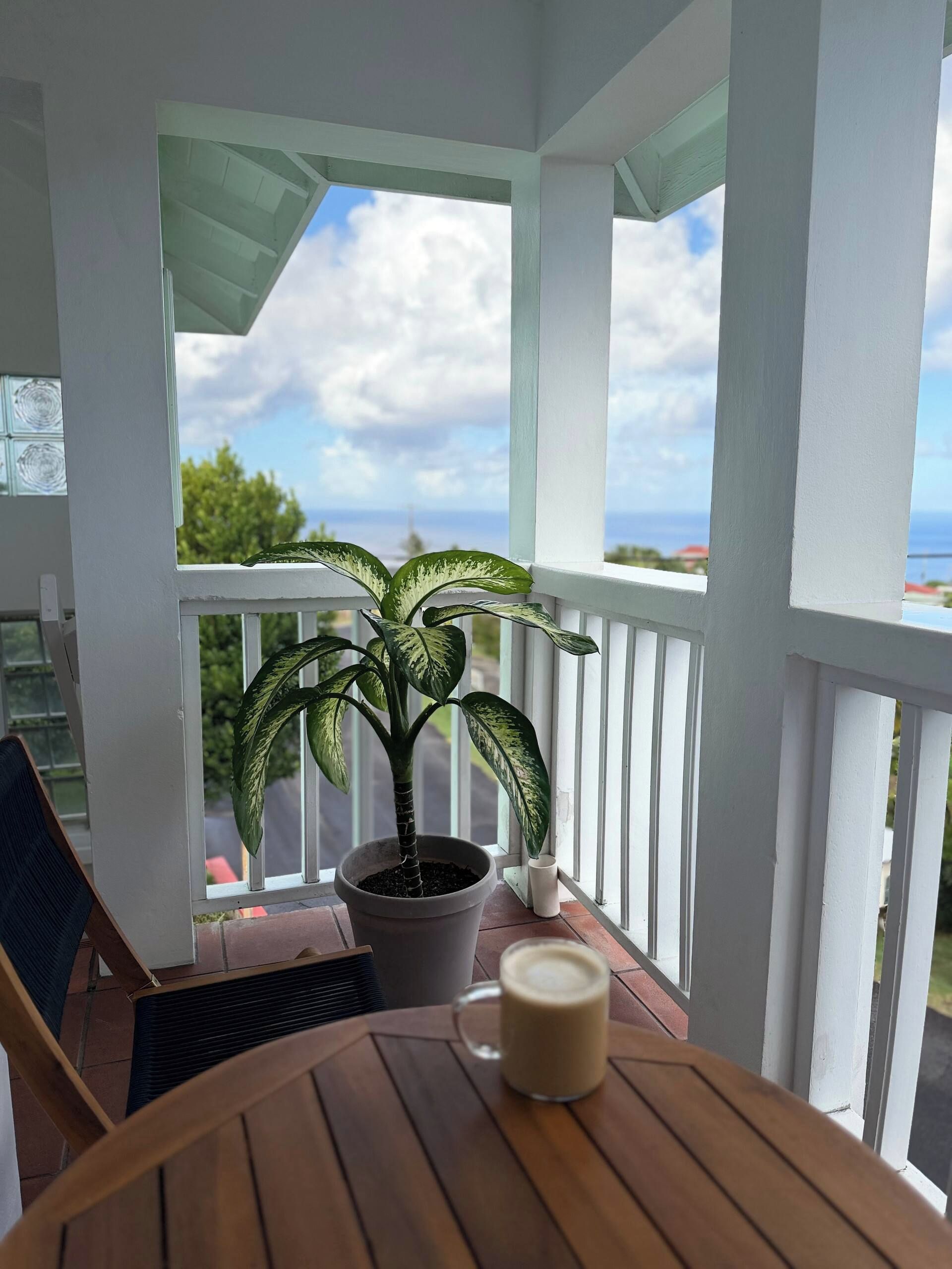 A balcony with a potted plant, a table with a mug, and a view of the sky and landscape.