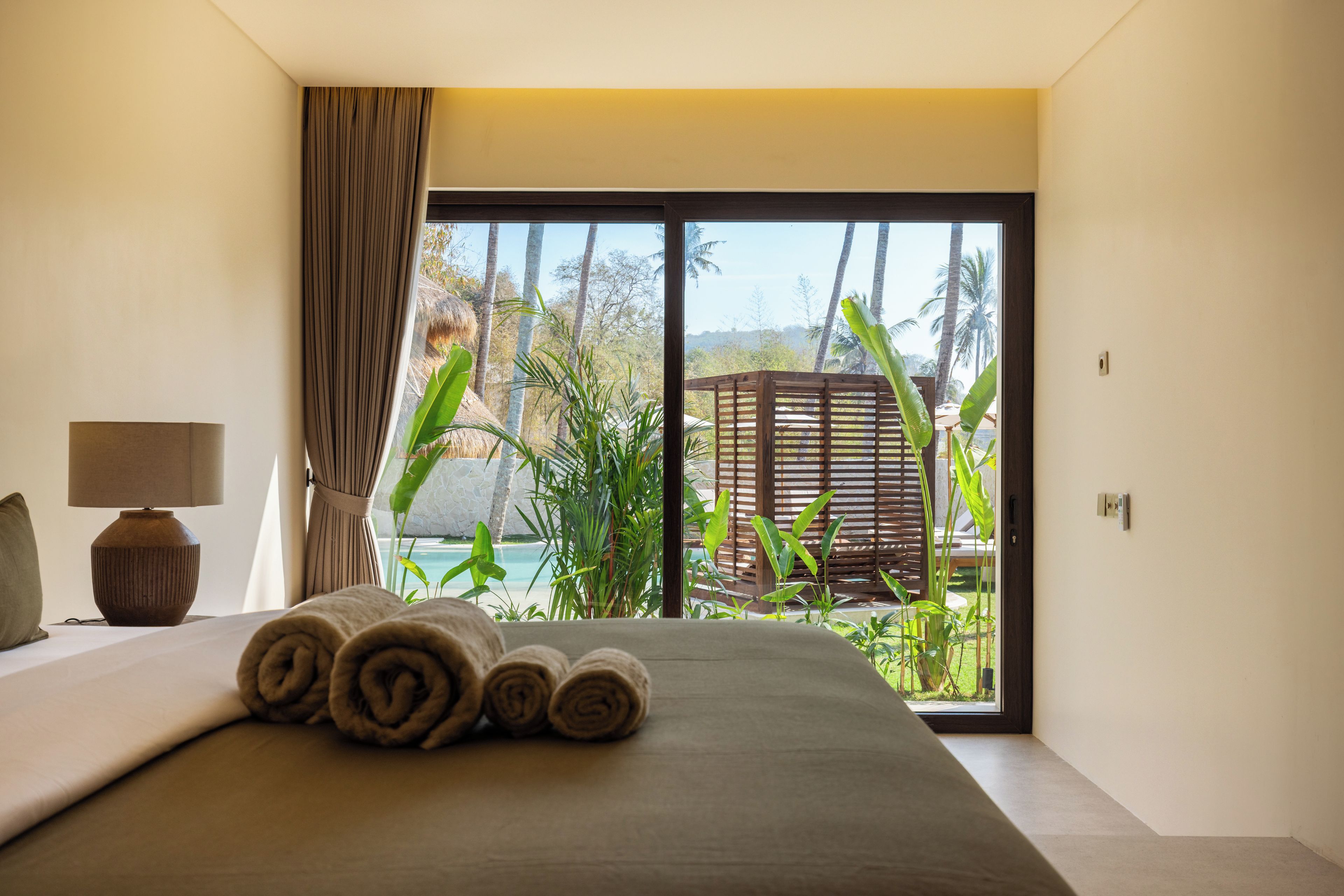 A hotel room with a bed, a lamp, and a view of a tropical landscape through a large window.
