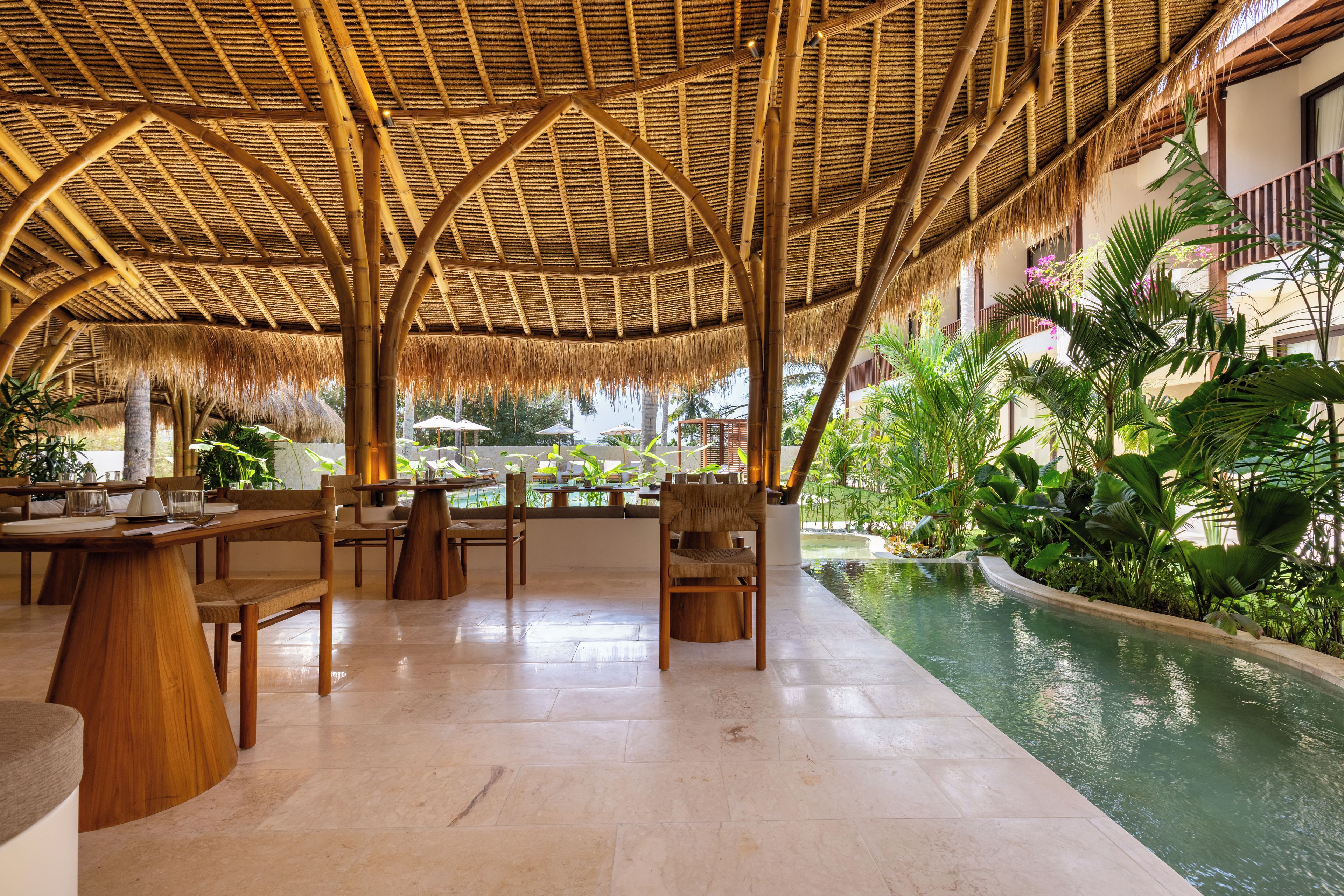 An indoor pool area with a thatched roof, wooden furniture, and tropical plants.
