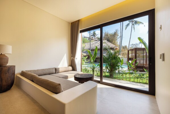 A modern living room with a sofa, a lamp, and a view of a tropical outdoor area with a pool.