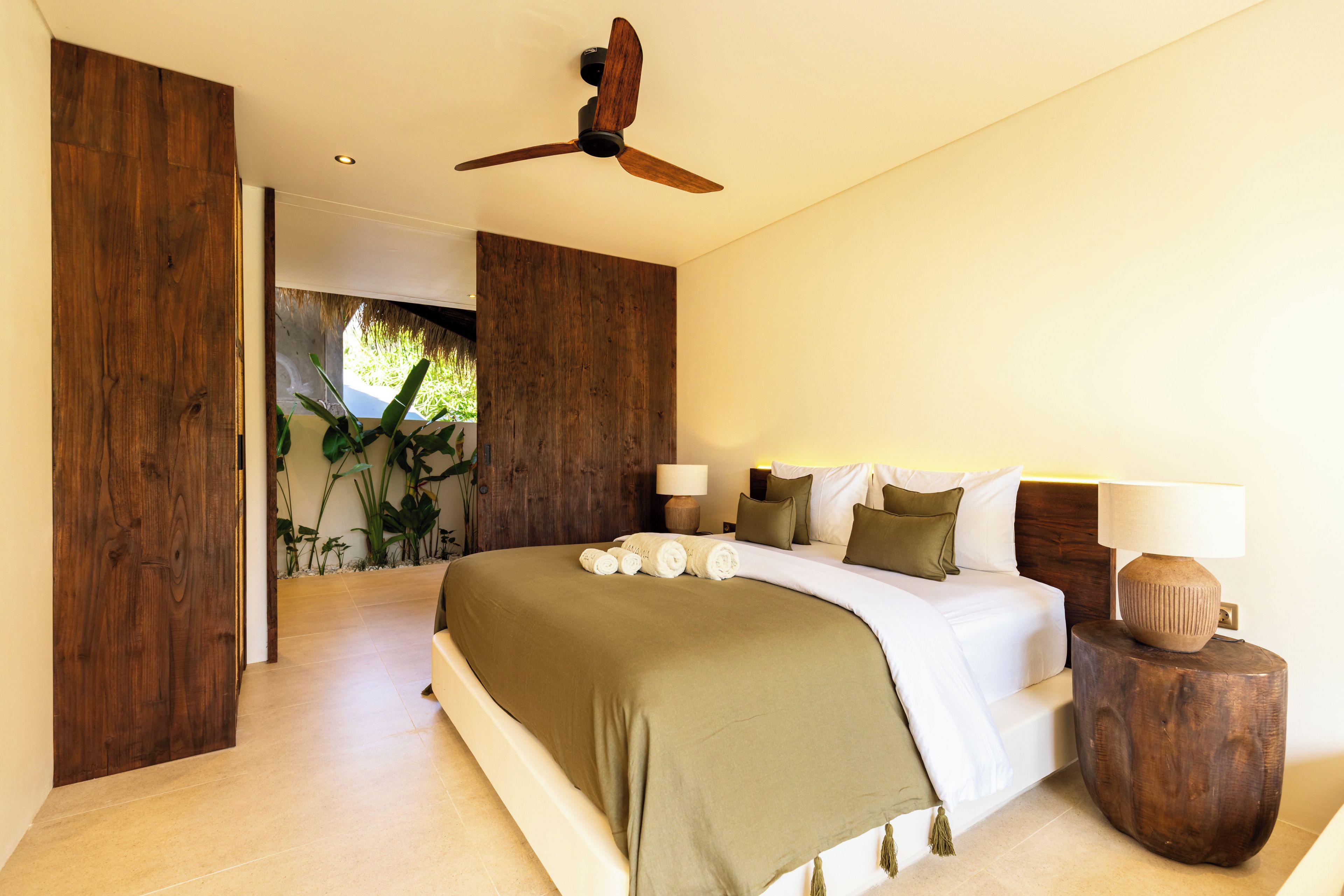 Quarto com cama grande, ventilador de teto e vista para um jardim tropical.