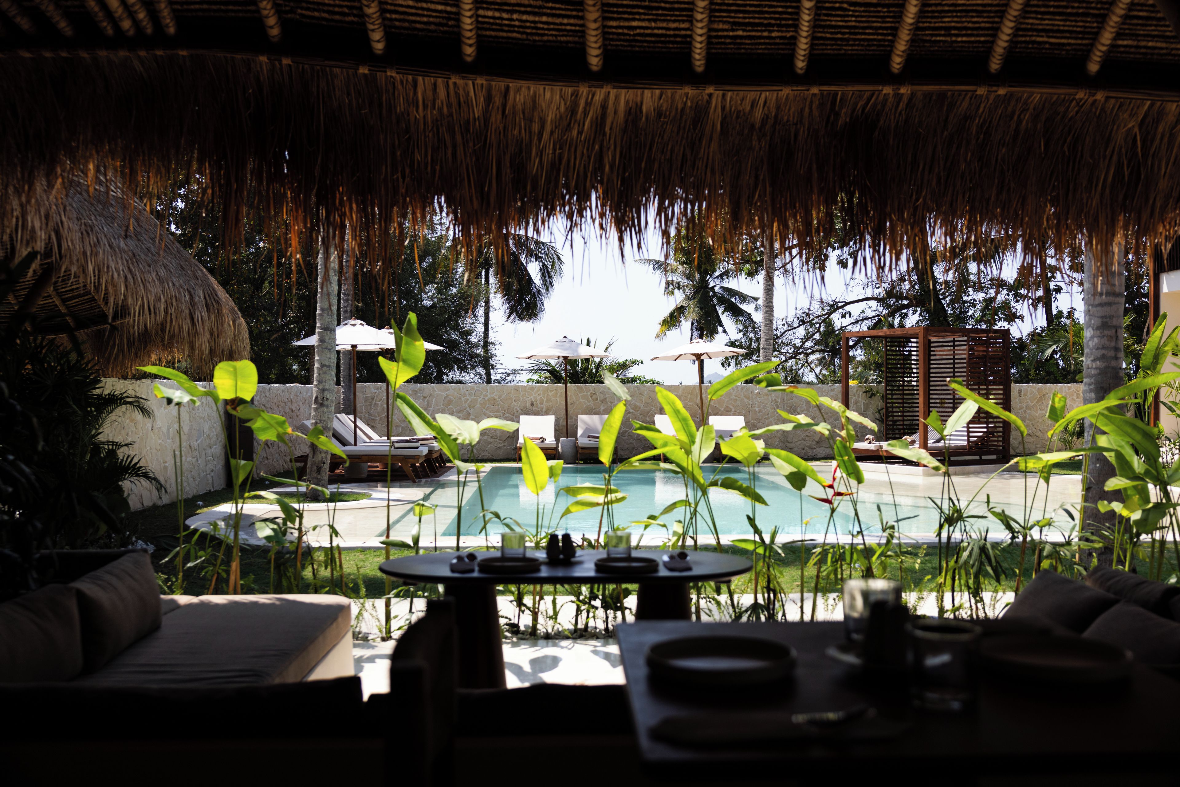 Uma área de piscina com espregui çadeiras, um telhado de palha e plantas tropicais.