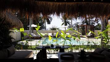 Uma área de piscina com espreguiçadeiras, um telhado de palha e plantas tropicais.