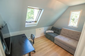 Ein kleiner, schräger Raum mit einem Schreibtisch, einem grauen Sofa und zwei Fenstern, die für natürliches Licht sorgen.