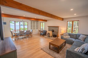 Ein großzügiges Wohnzimmer mit Kamin, einer Couch, einem Essbereich mit Stühlen und großen Fenstern mit Blick ins Freie.