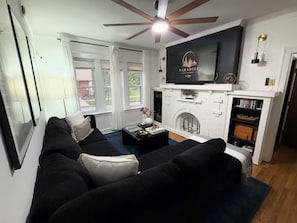 A modern living room with a black sofa, a flat-screen TV mounted above a fireplace, and a ceiling fan.