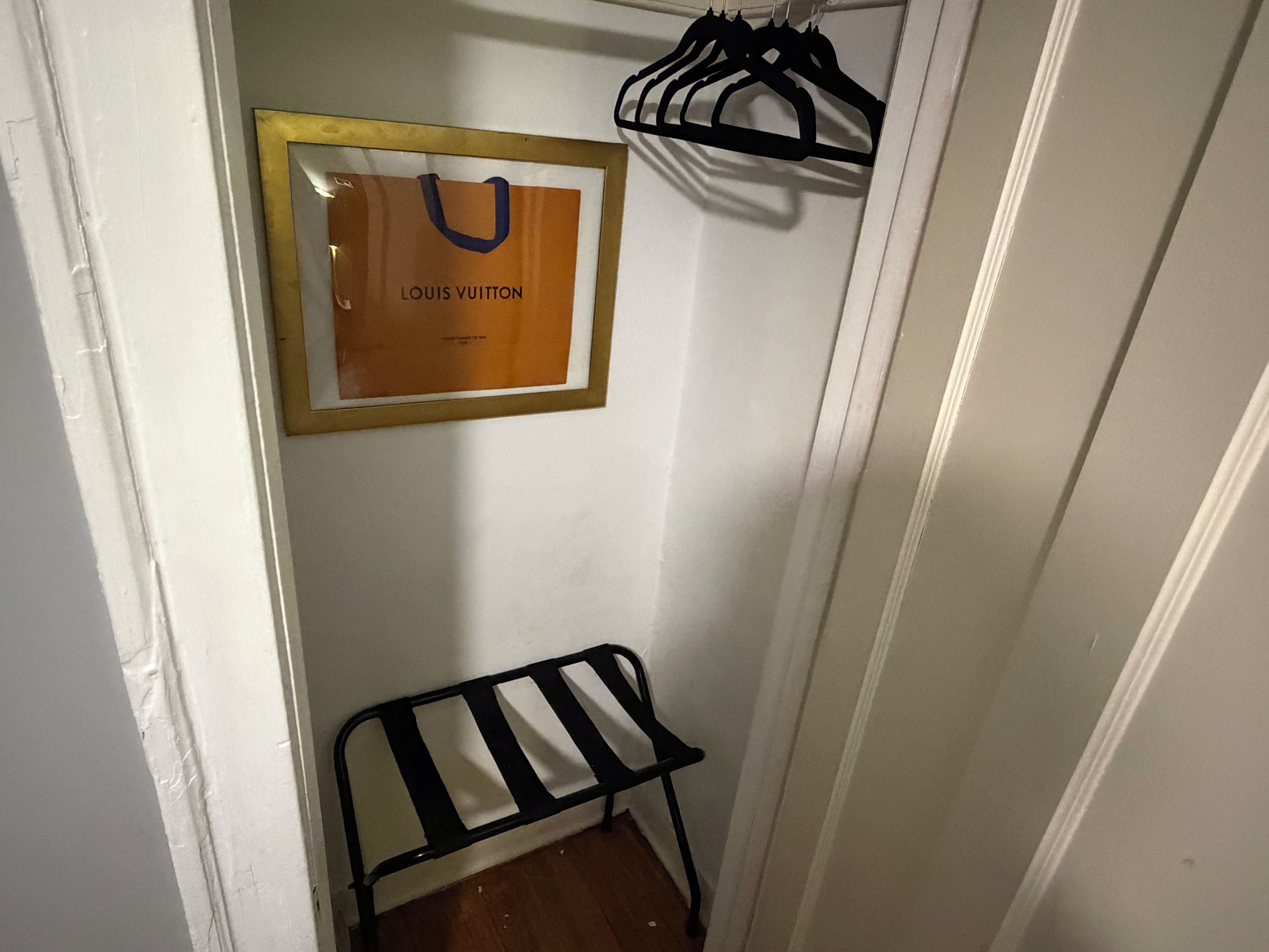 A narrow hallway with a framed Louis Vuitton bag on the wall, a black metal chair, and a black hanger rack.
