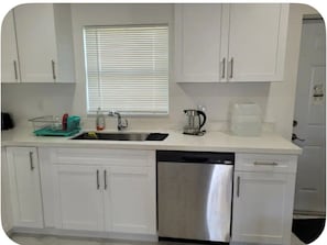 A kitchen with white cabinets, a stainless steel dishwasher, a kettle, and a coffee maker. - Sun‑Soaked Living in the Heart of Florid, 3‑Bd House Available in Lehigh Acres! (Lehigh Acres)