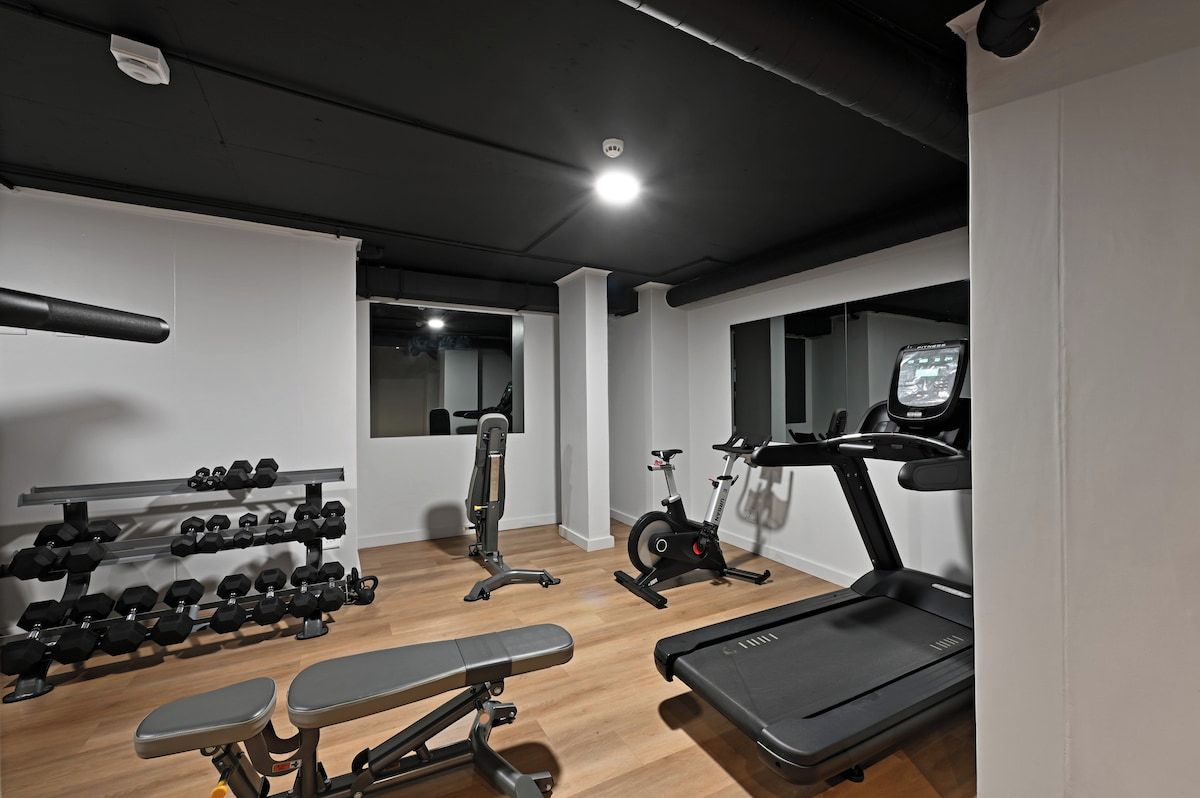 Een fitnessruimte met trainingsapparatuur, waaronder gewichten, een bankje, een hometrainer en een loopband.