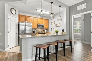 Eine moderne Küche mit einem Edelstahl-Kühlschrank, Holzschränken, einer Mikrowelle und einem eingebauten Backofen. Drei Barhocker und Pendelleuchten befinden sich über der Kochinsel.