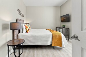 A bedroom with a bed, a bedside table, a lamp, a painting on the wall, and a door. - Orlando 3BR Condo, Space, Comfort and Location (Orlando)