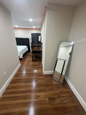 A narrow hallway with wooden flooring, a bed with a white cover, a dark-colored wardrobe, and a mirror leaning against the wall.