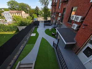 A residential area with a paved pathway, green artificial grass, and a brick building with air conditioning units.
