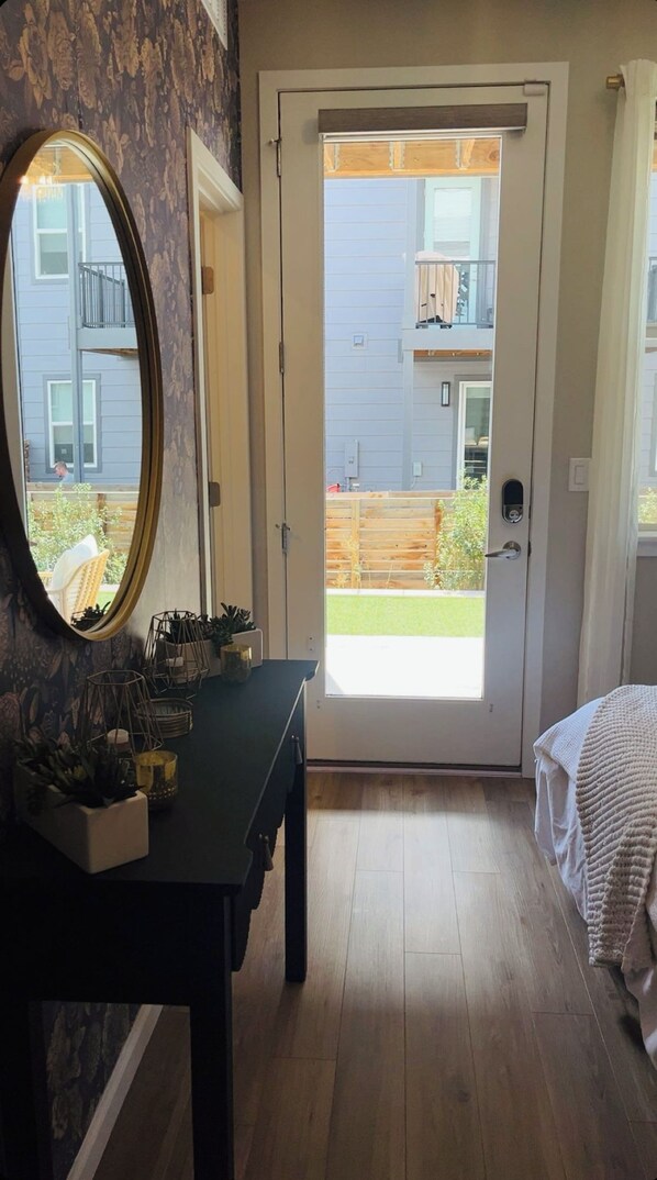 A room with a wooden floor, a bed with a white blanket, a black dresser with a mirror, and a door leading to a balcony with a view of a building and a fence.