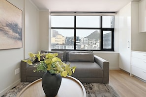 Un salon moderne avec un canapé gris, une table ronde en bois surmontée d’un vase noir, et une grande fenêtre donnant sur la ville.