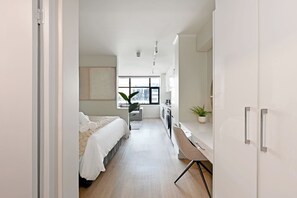 Un dormitorio moderno y luminoso con un ventanal grande, cama con ropa de cama blanca, una pequeña zona de estar y una kitchenette con lavadora.