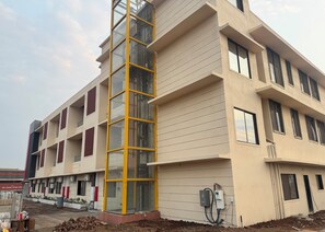 A multi-story building with a central glass elevator shaft, multiple windows, and a visible electrical box on the ground level. - Hotel Food Avenue (Bhiwandi)