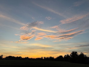 A sunset over a rural landscape with a clear sky and silhouettes of trees and houses.