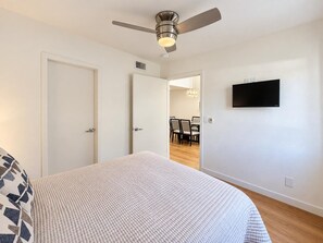Ein Schlafzimmer mit einem Bett, einem Deckenventilator, einem an der Wand montierten Fernseher und einer Tür zu einem Essbereich.
