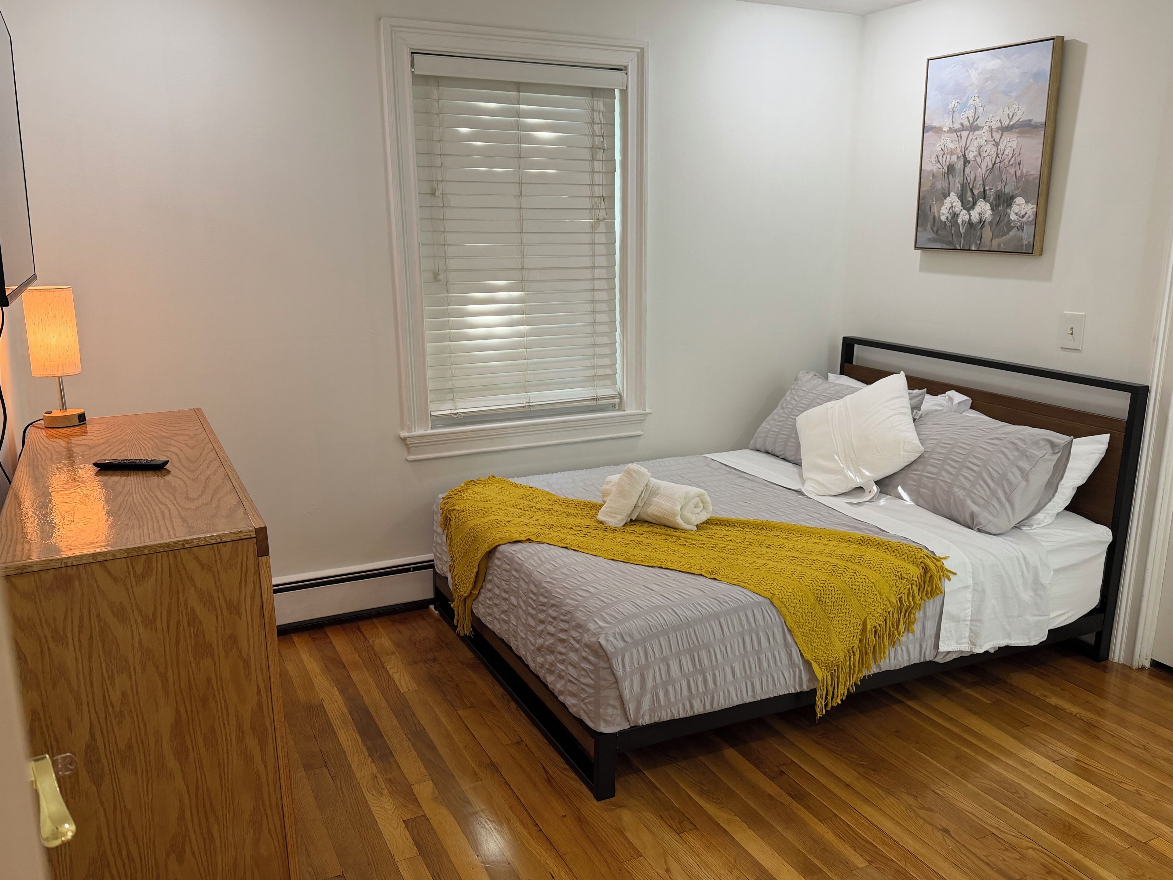 A bedroom with a bed, a bedside table, a lamp, a window with blinds, and a painting on the wall.
