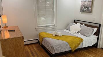 A bedroom with a bed, a bedside table, a lamp, a window with blinds, and a painting on the wall.