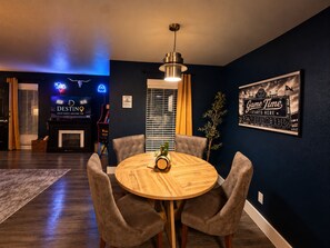 Ein Zimmer mit einem runden Holztisch, vier gepolsterten Stühlen, einer Deckenleuchte, einem gerahmten Bild an der Wand und einem Neonzeichen.