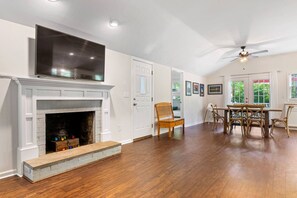 A spacious room with hardwood flooring, a fireplace, a dining area with a table and chairs, and a ceiling fan.