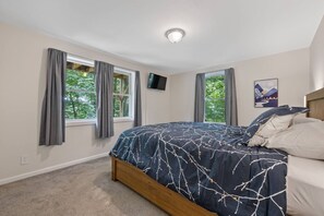 A bedroom with a bed, two windows with curtains, a flat-screen TV on the wall, and a framed picture.