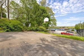 A basketball court with a hoop, surrounded by trees and a parking area with cars. - Hilltop Hideaway · Sleeps 9 · Near Lake Hayward & Mohegan Sun (Colchester)