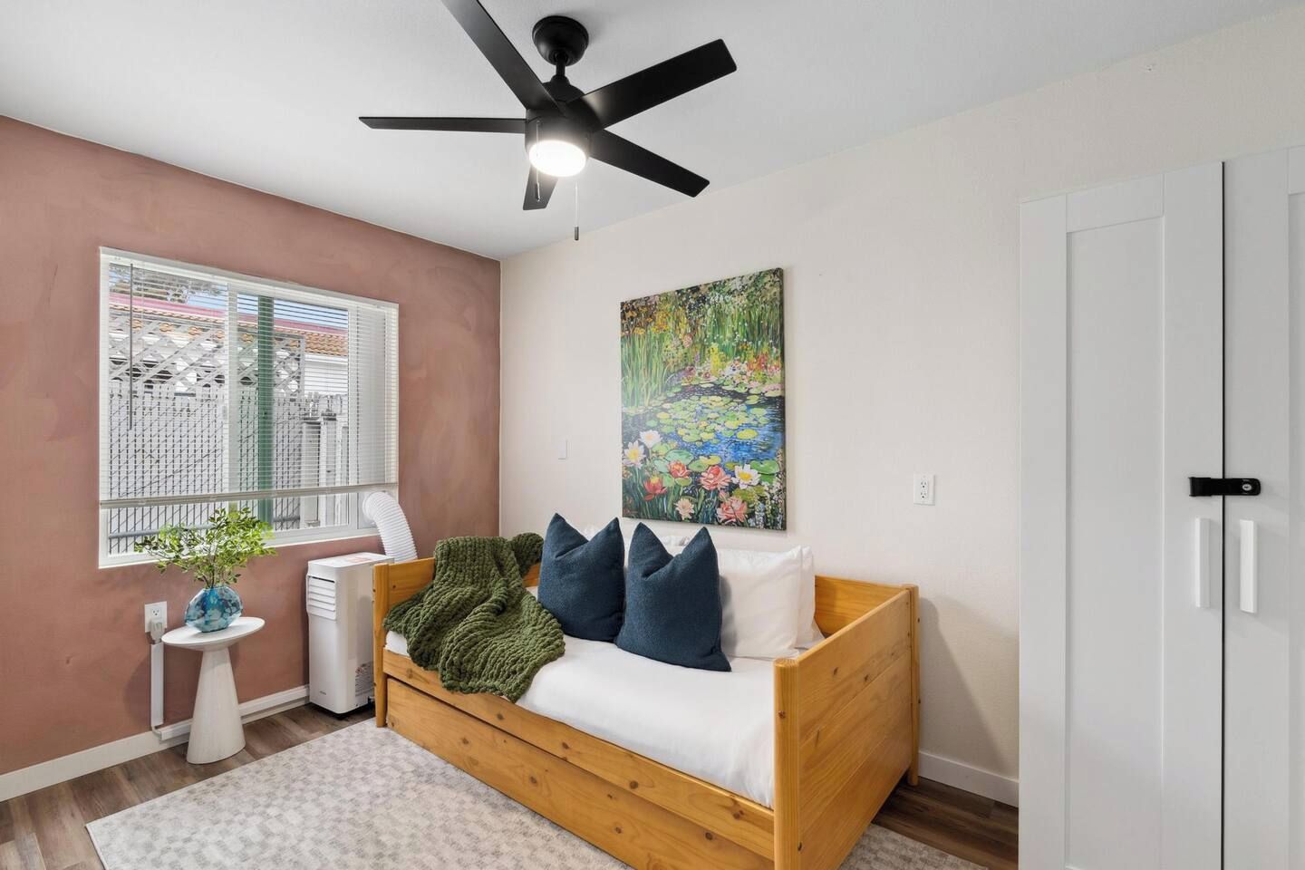 A room with a bed, a ceiling fan, a window with blinds, a radiator, a painting, and a door.