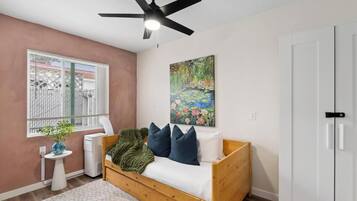 A room with a bed, a ceiling fan, a window with blinds, a radiator, a painting, and a door.