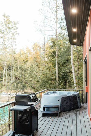 Une terrasse en bois avec un jacuzzi et un barbecue, entourée d’arbres.