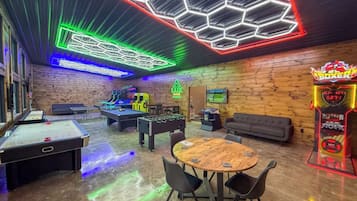 A room with a pool table, foosball table, and a table with chairs. There is a neon sign on the wall and a video game machine.