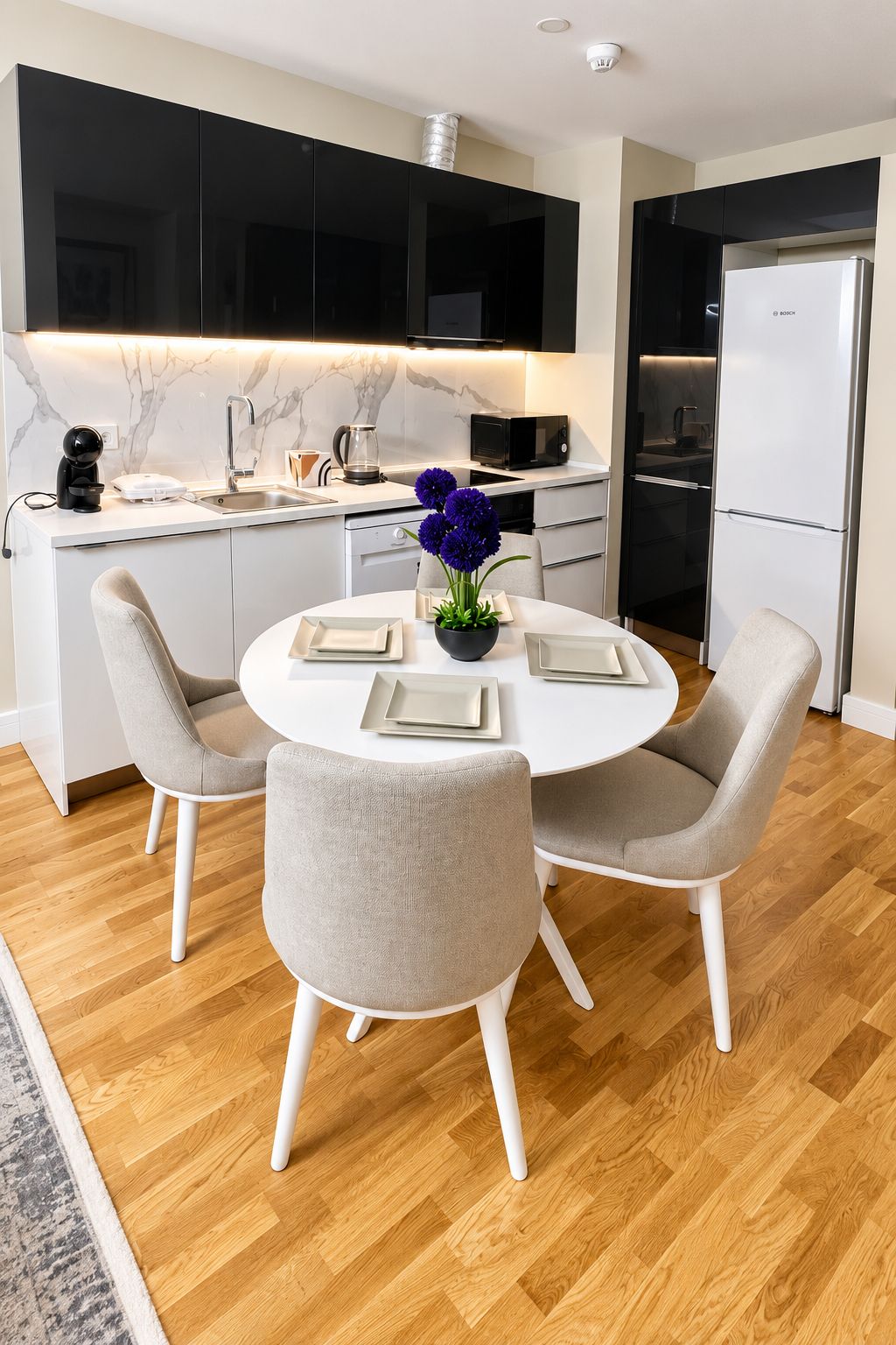 A modern kitchen with a dining area, featuring a round table with chairs, a refrigerator, and a microwave.