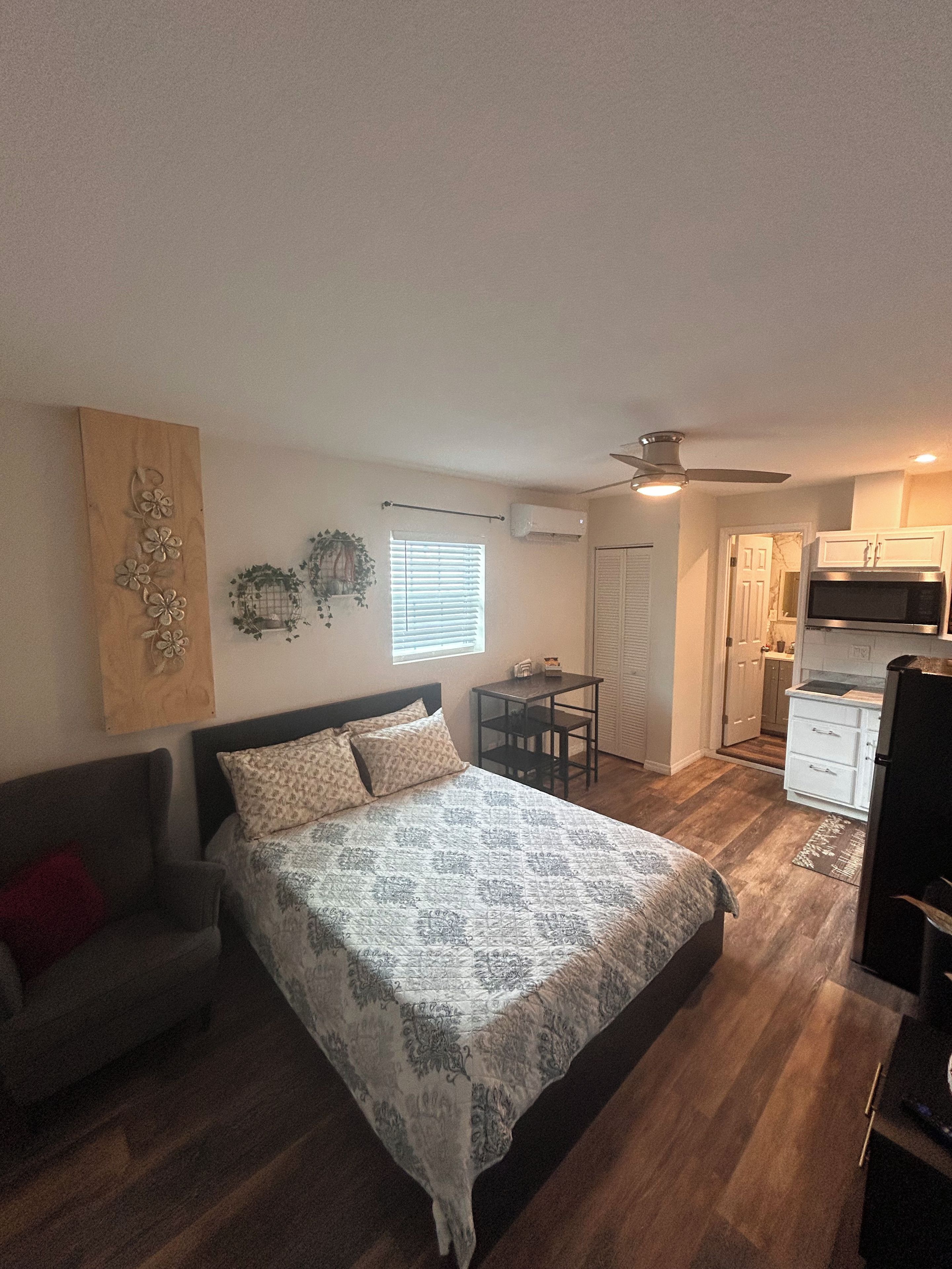 A bedroom with a bed, a small table, and a sofa. There is a kitchenette with a microwave and a refrigerator.