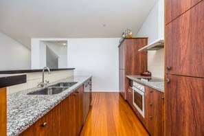 A compact kitchen with granite countertops, a stainless steel sink, wooden cabinets, and a built-in oven. - Dual-Level Harbour Apartment on The Wharf (Auckland City)