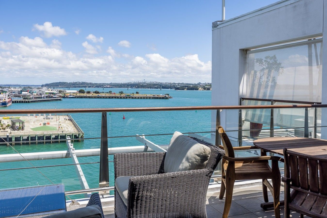 A balcony with wicker furniture, a wooden table, and a view of a harbor with boats and a pier.
